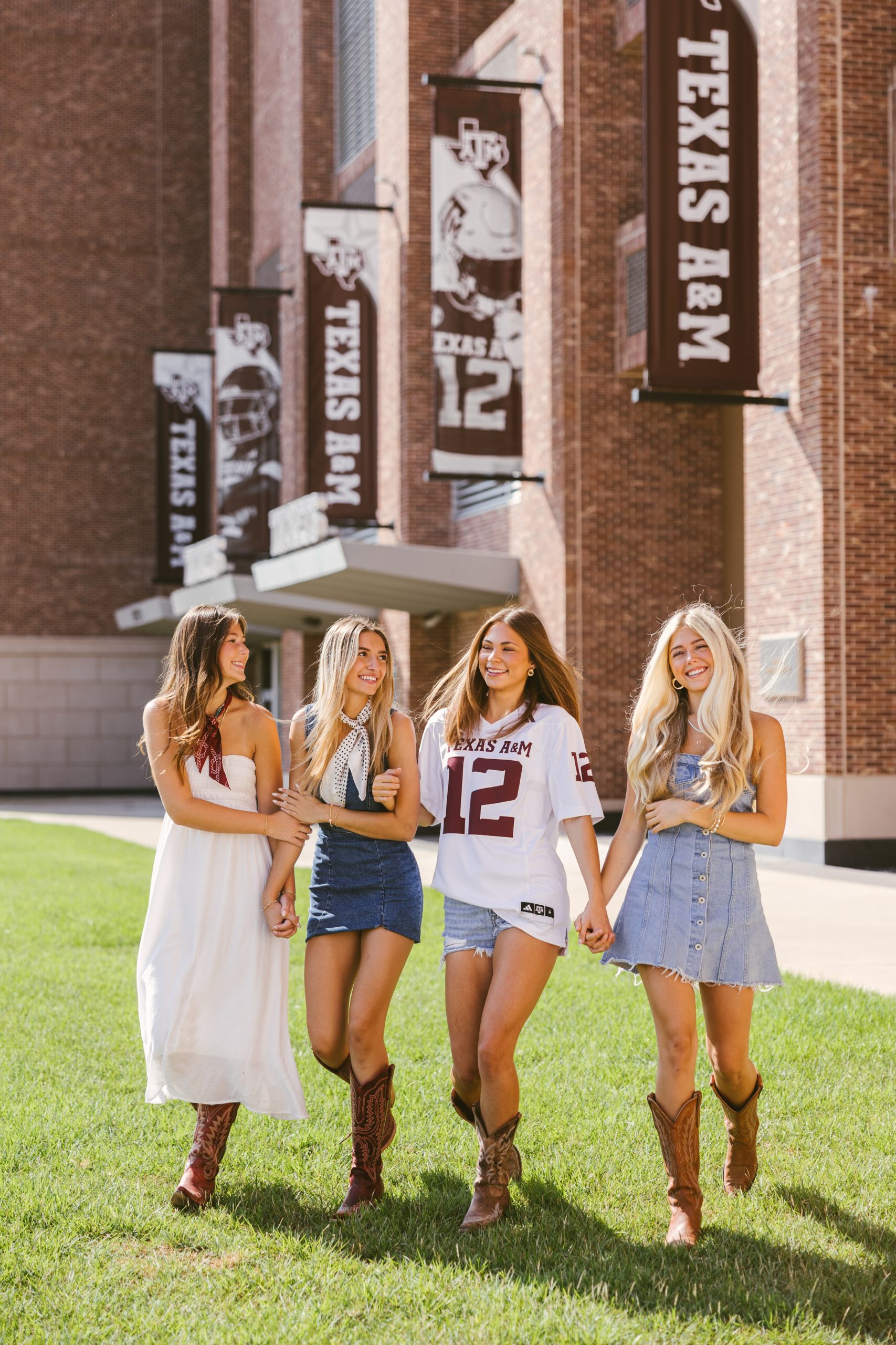 Four young women wearing casual and western-inspired outfits, including cowboy boots, walk together and smile outside a Texas A&M University building with large banners displaying the school’s name and logo.