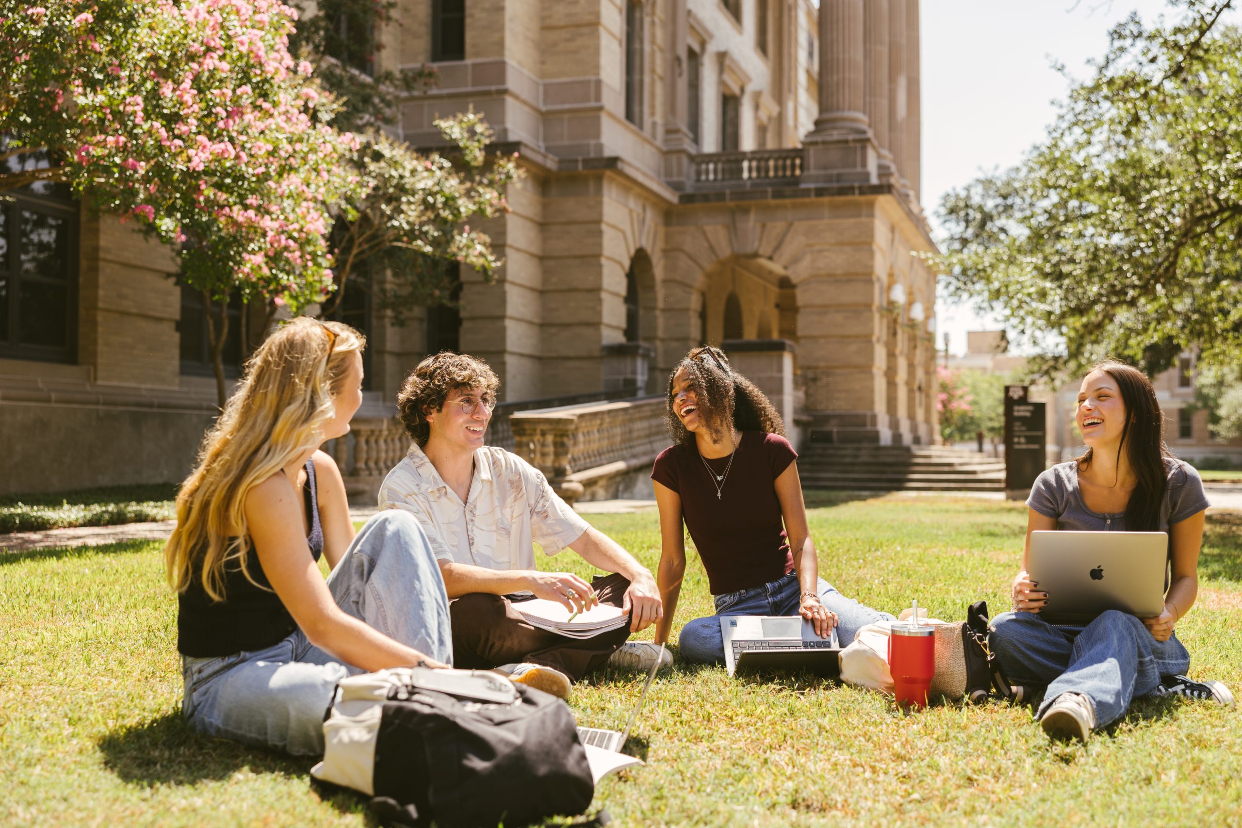 Four college students sit on the grass outside a campus building, smiling and talking. Two have laptops, and one has a drink container. Trees and sunlight create a bright, cheerful atmosphere.