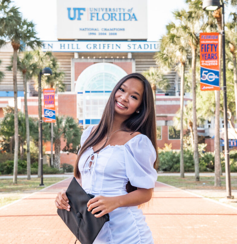 A young woman in a white dress smiles and holds a black folder while standing on a brick walkway in front of the University of Florida stadium, with palm trees and celebratory banners in the background.