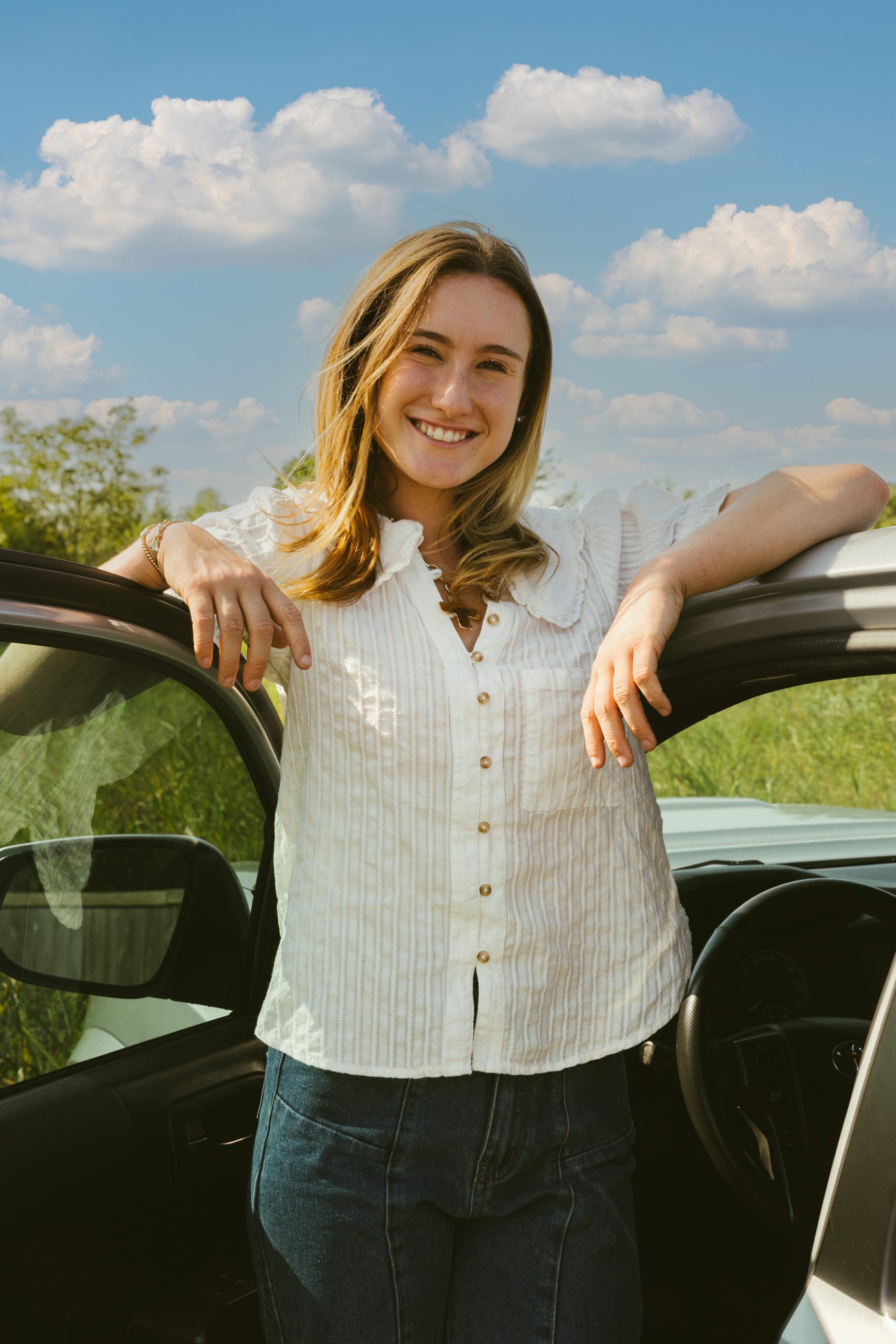 A young woman with long hair, wearing a white blouse and jeans, smiles while standing outside by an open car door on a sunny day, with a background of green grass and a blue sky with clouds.