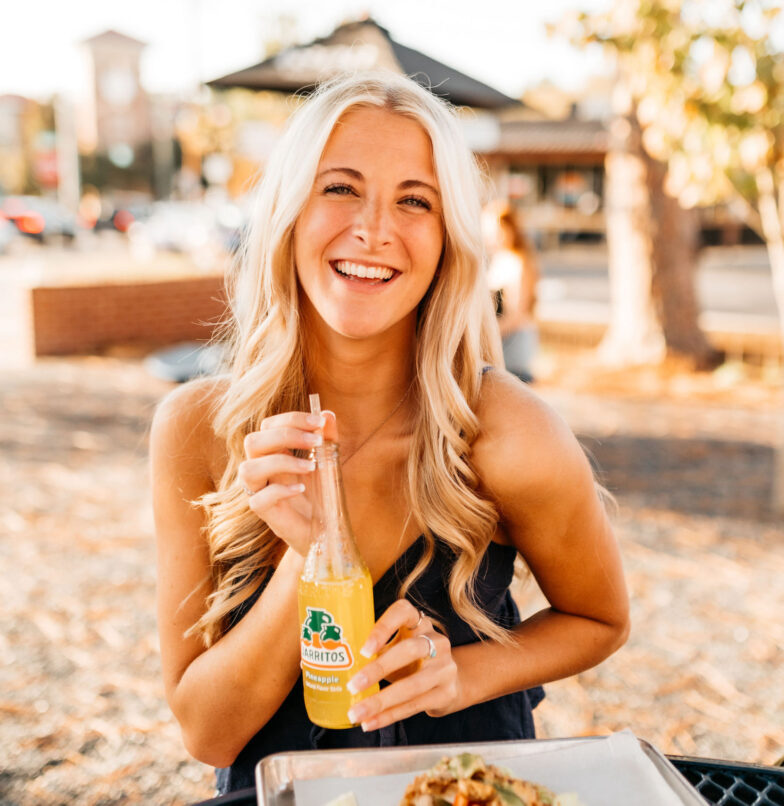 A smiling woman with long blonde hair sits outdoors, holding a glass bottle of Jarritos pineapple soda and looking at the camera. There is a tray of food in front of her and sunlight filters through the trees in the background.