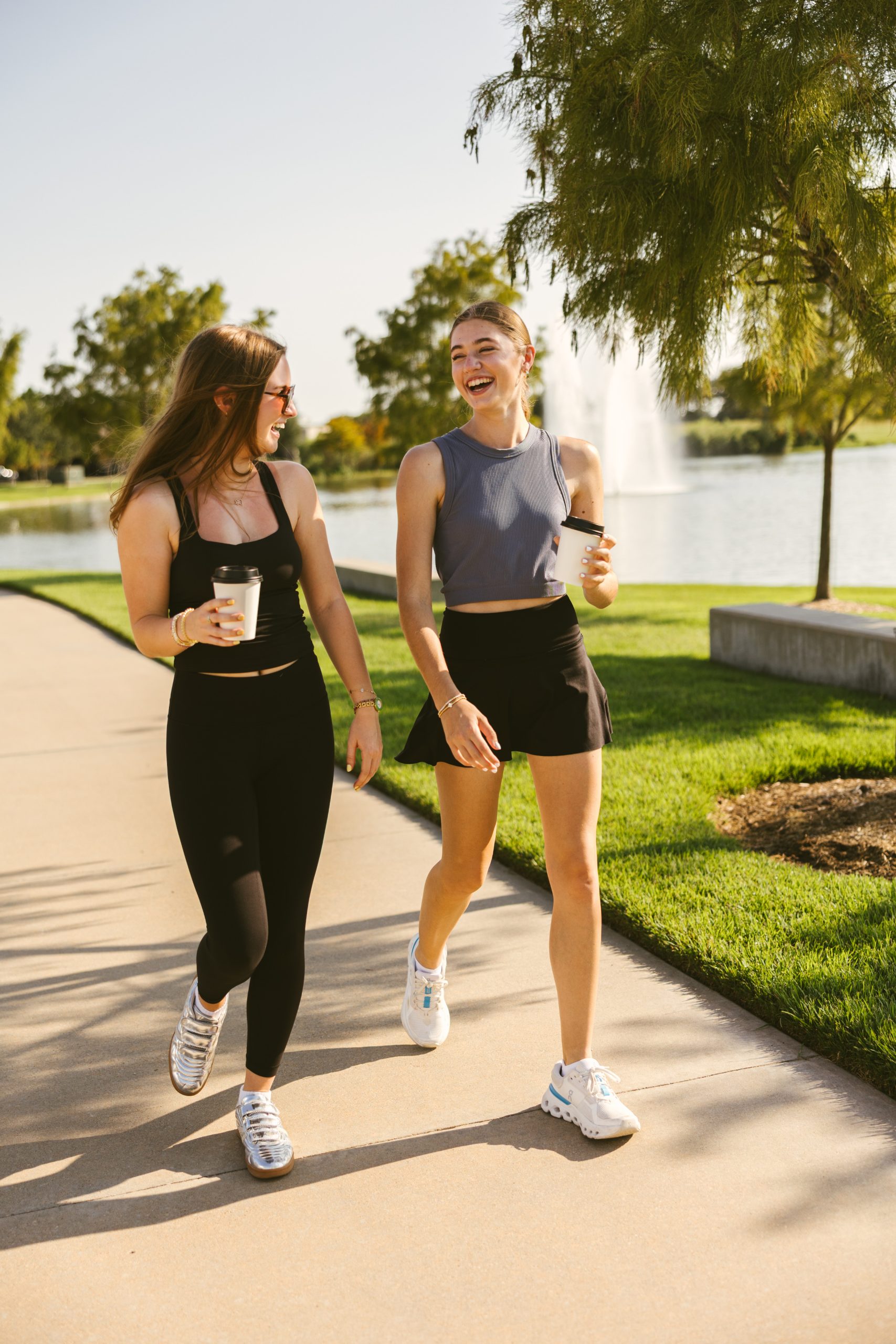 Two women in athletic wear walk on a sunny path by a lake, holding coffee cups, smiling and chatting. A fountain sprays water in the background, and trees and grass surround the area.