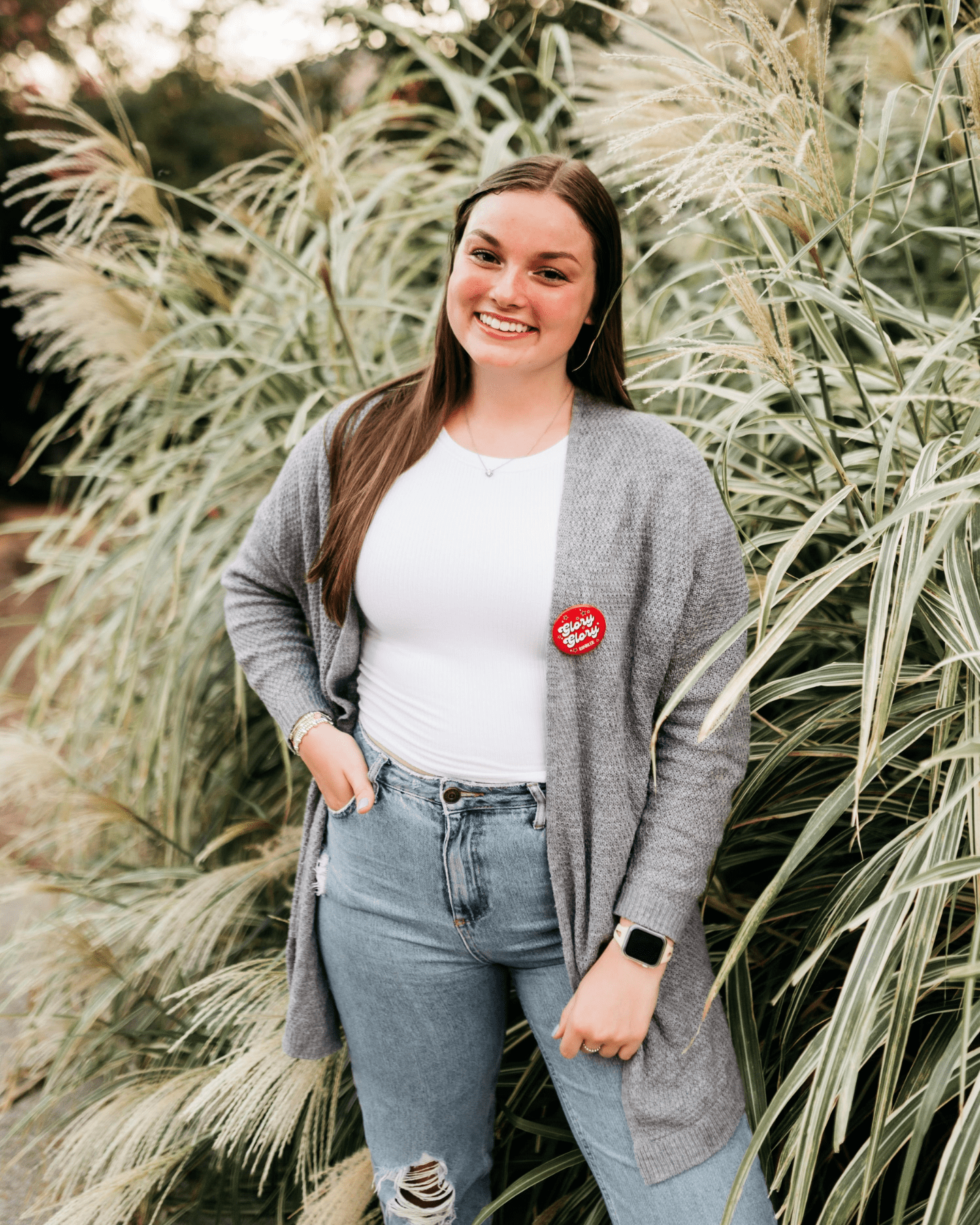 A young woman with long brown hair stands smiling in front of tall ornamental grasses, wearing a gray cardigan, white top, ripped jeans, and a red button that says Senior 2023.