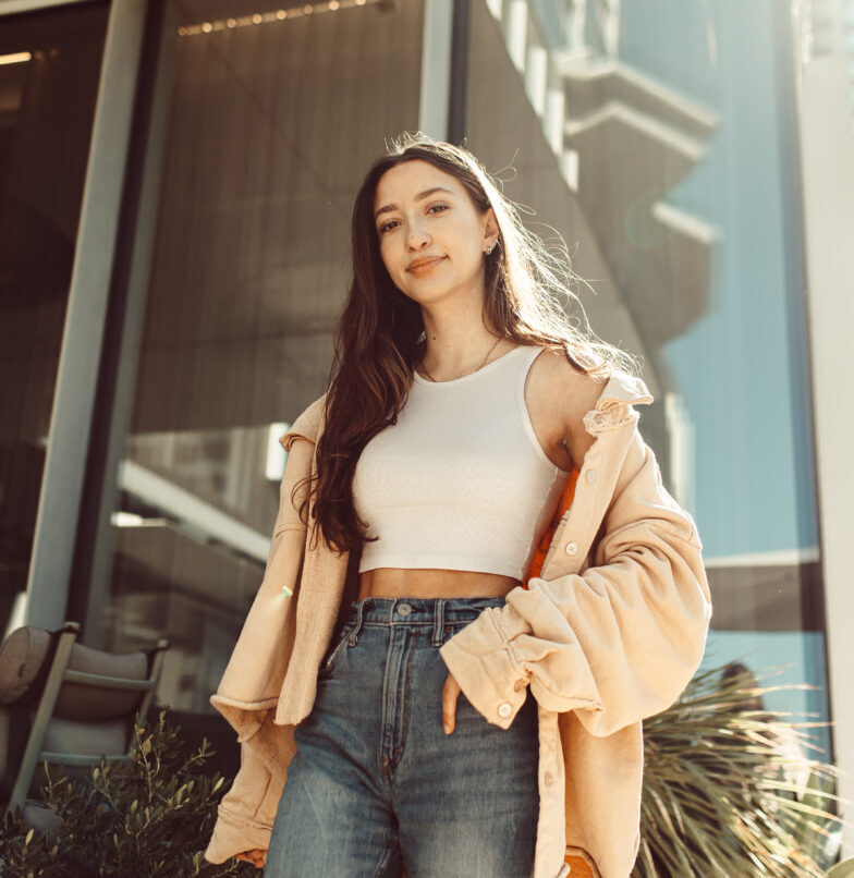 A young woman with long brown hair stands confidently outside a modern building, wearing a beige jacket over a white crop top and high-waisted jeans, with sunlight shining on her.