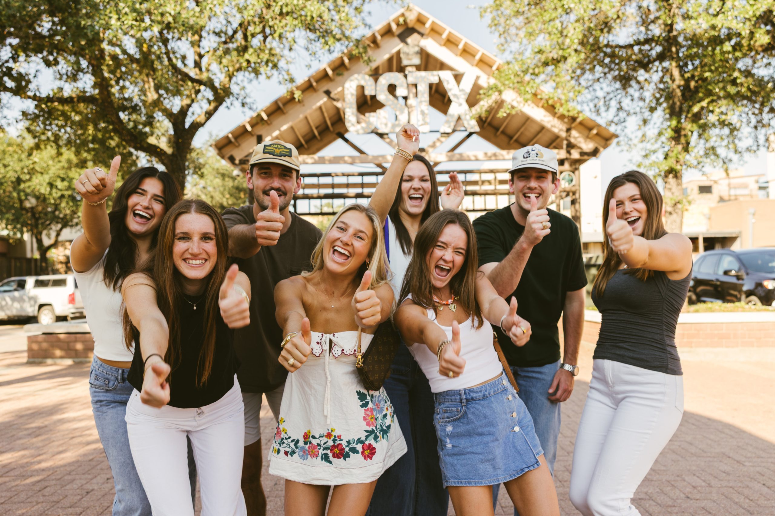 A group of smiling young adults stand outdoors in front of a building with a CSTX sign, giving thumbs up and looking cheerful, surrounded by trees and sunlight.