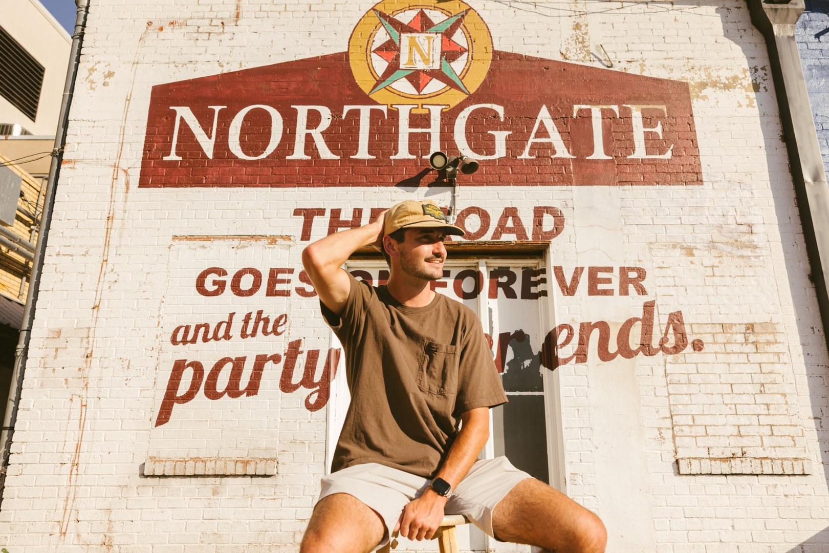 A man in a tan hat and brown shirt sits in front of a brick wall painted with NORTHGATE and The road goes on forever and the party never ends, enjoying the sunny day near apartments near Texas A&M. He is smiling and holding his hat.
