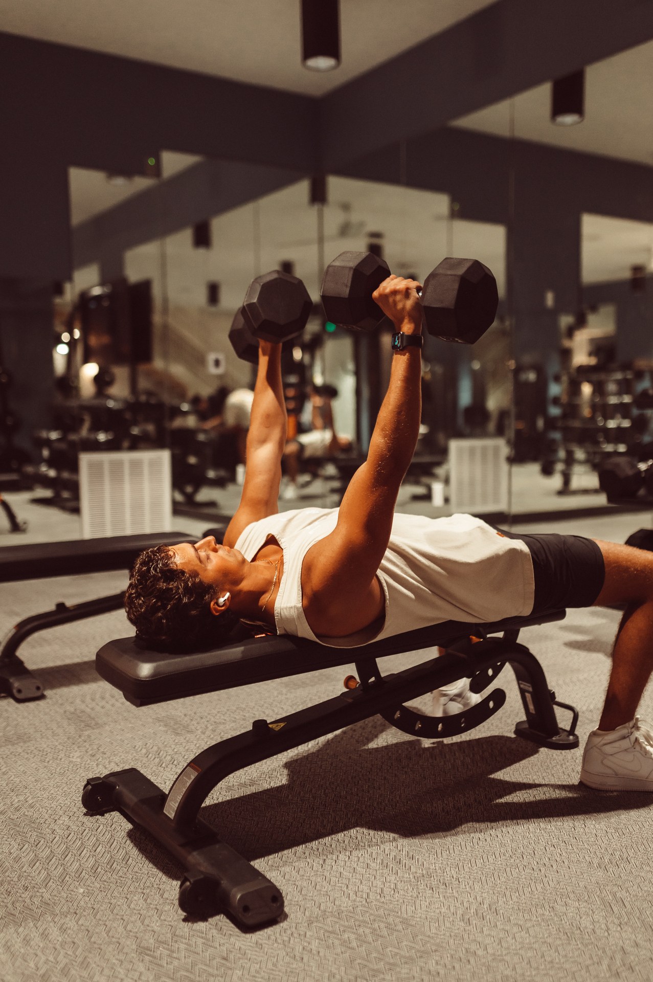A person in a white tank top and black shorts performs a dumbbell bench press on a workout bench in a gym, possibly located in one of the off campus apartments TAMU students prefer, with large mirrors and gym equipment visible in the background.