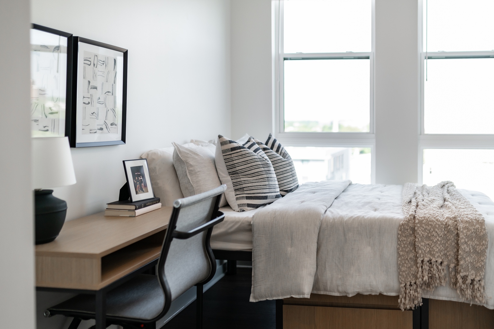 A modern, minimal bedroom in off campus apartments TAMU, featuring a neatly made bed with neutral bedding and striped pillows, a cozy throw, a tidy desk with lamp and books, and large windows that flood the space with natural light.
