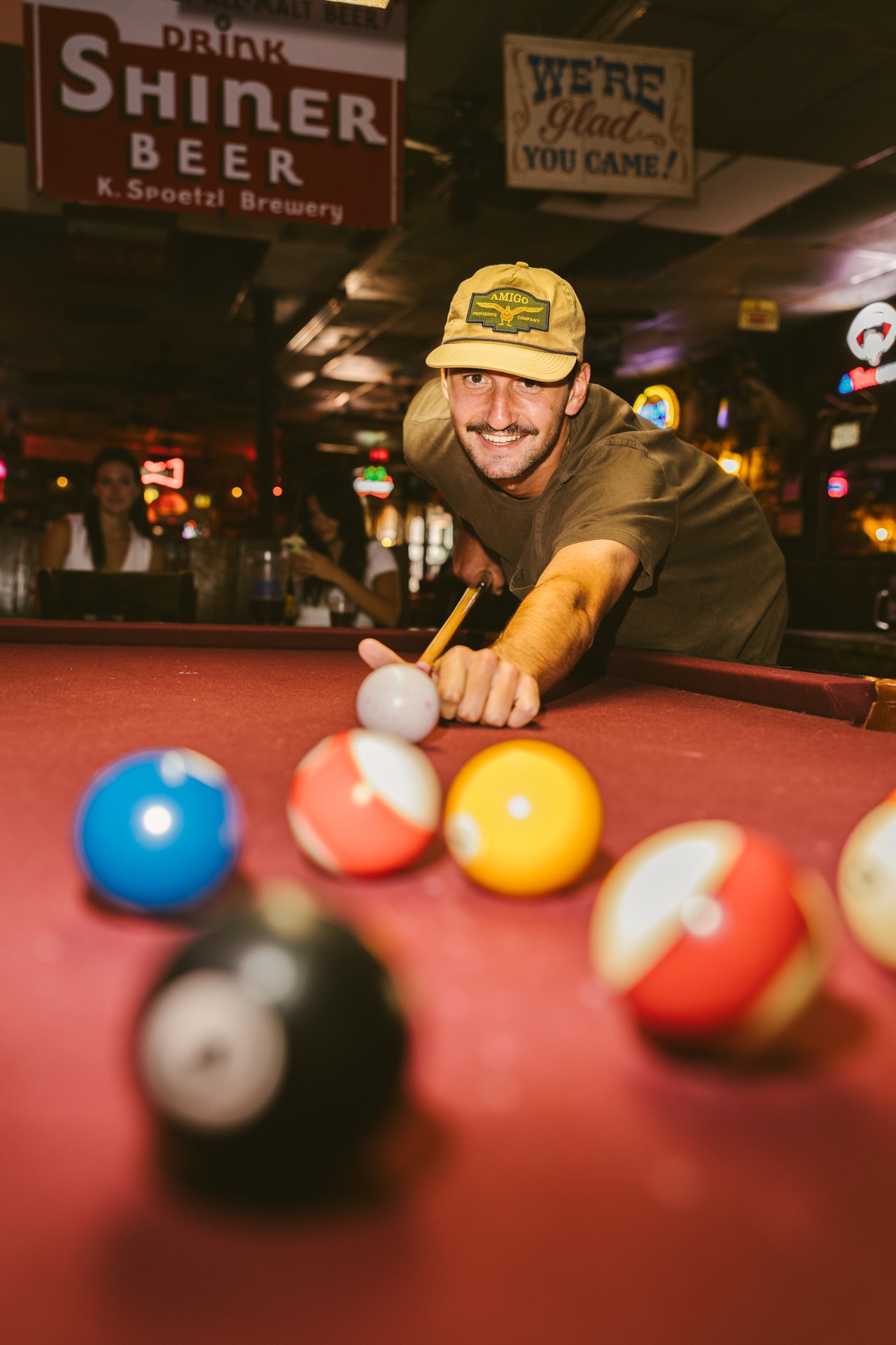 A man in a beige cap smiles as he lines up a shot on a red pool table in a dimly lit bar near off campus apartments TAMU, with colorful billiard balls in the foreground and bar signs glowing in the background.