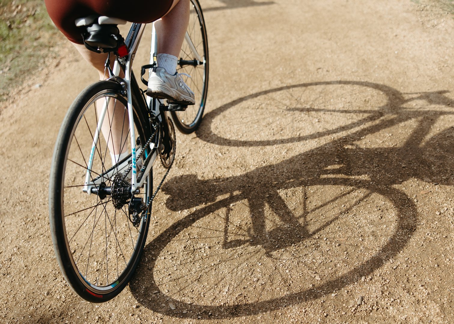 Close-up of a person riding a bicycle near off campus apartments TAMU on a dirt path, showing the back wheel, part of the frame, and their legs. The bike and legs cast a clear shadow on the ground.