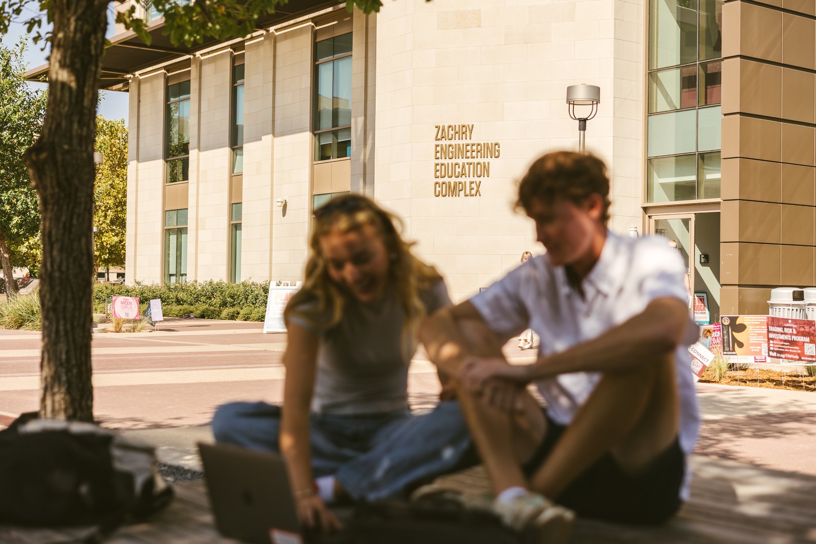 Two students sit on the ground under a tree, working on a laptop outside the Zachry Engineering Education Complex building on a sunny day, enjoying campus life close to popular apartments near Texas A&M University.