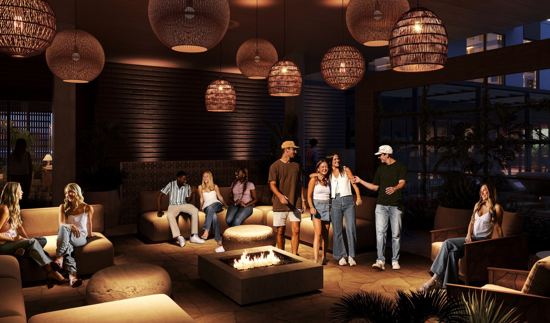A group of young adults relax and socialize around a fire pit at The Jay, a cozy, dimly lit lounge with warm lighting and natural decor. Some sit on sofas while others stand and chat, creating a friendly, inviting atmosphere.