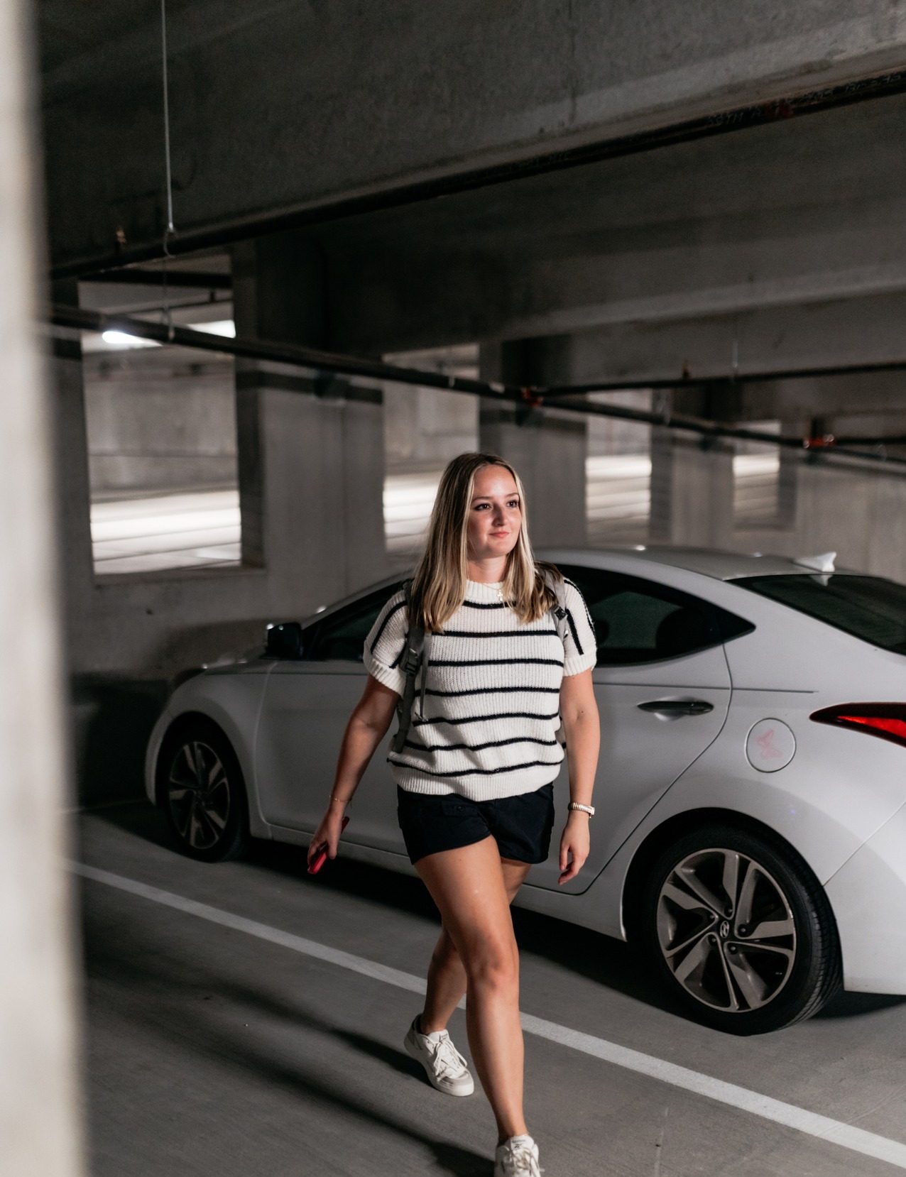 A woman in a white and black striped top, black shorts, and white sneakers walks through a parking garage beside a white car, holding her phone—perhaps checking directions to off campus apartments TAMU students prefer.