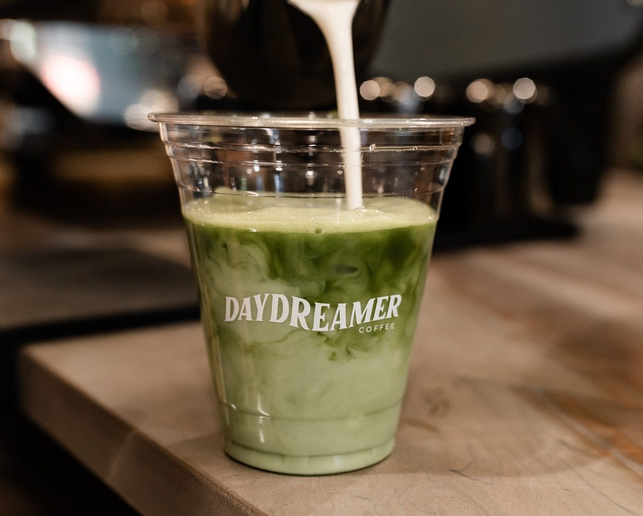 A hand pours milk from a metal pitcher into a plastic cup of iced matcha tea with Daydreamer Coffee printed on the cup, set on a wooden counter near off campus apartments TAMU.