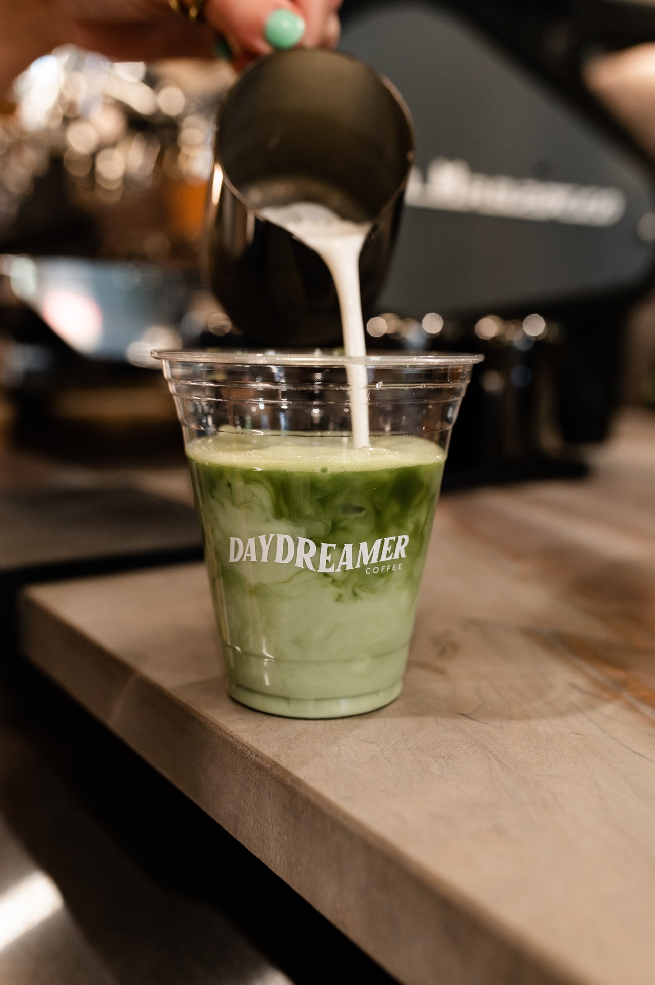 A hand pours milk from a metal pitcher into a plastic cup of iced matcha tea with Daydreamer Coffee printed on the cup, set on a wooden counter near off campus apartments TAMU.