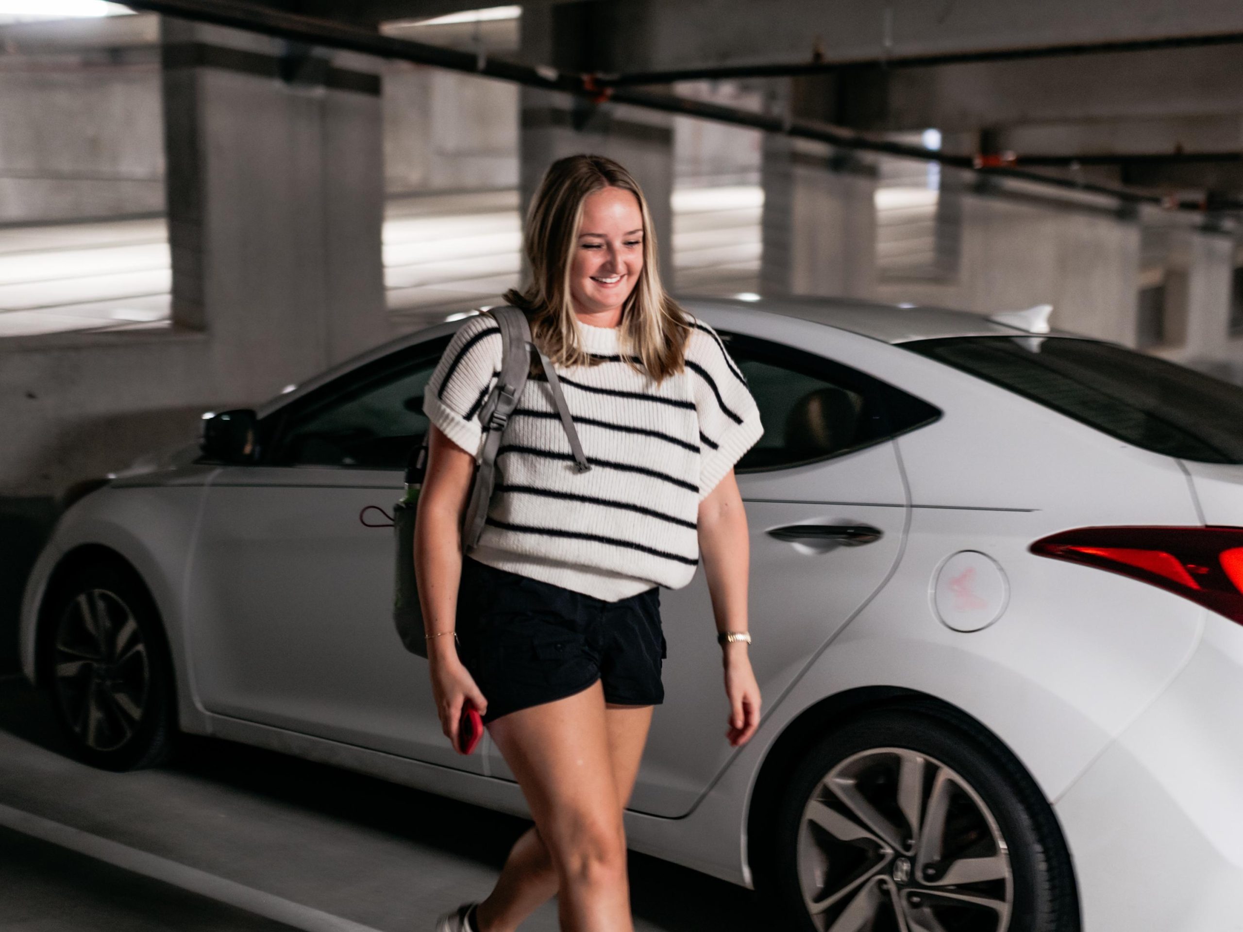A woman with shoulder-length hair, wearing a white and black striped shirt and black shorts, walks in a parking garage next to a white car, smiling and holding a red phone in her hand.