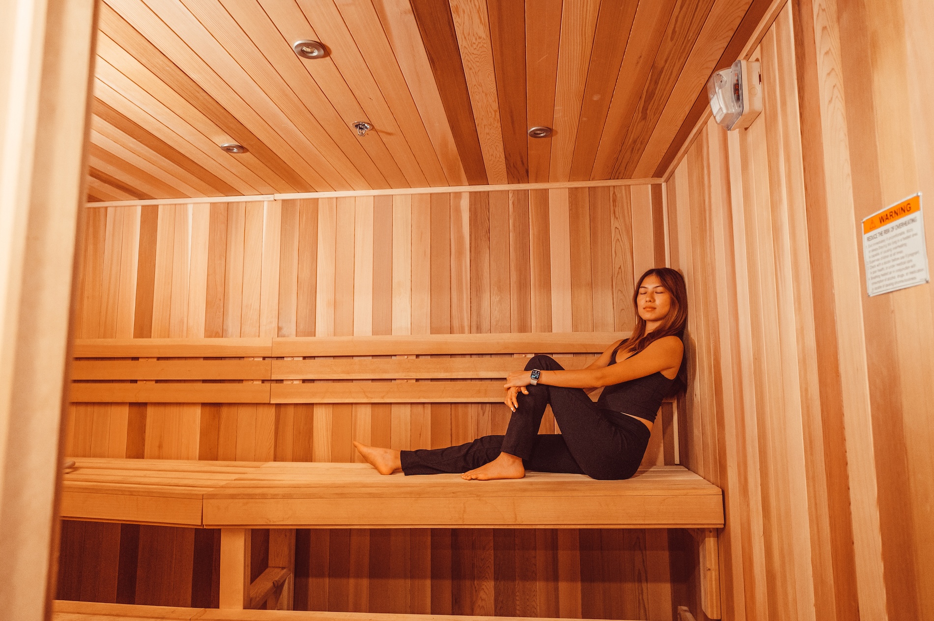 A woman sits barefoot on the top wooden bench inside a modern, empty sauna room, leaning back with one arm resting on her knee. The walls and ceiling are lined with light-colored wood panels.