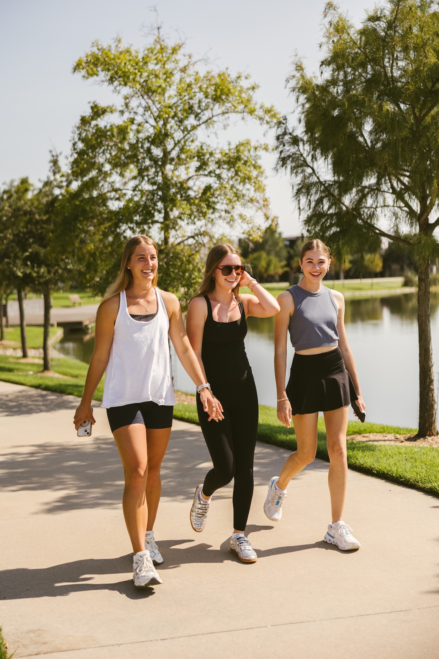 Three young women in athletic outfits walk and smile together on a sunny path beside a lake, surrounded by trees and greenery near apartments near Texas A&M University.