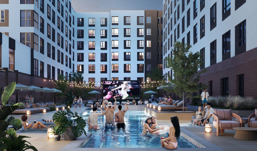 People relax and socialize around a modern outdoor swimming pool at dusk, with apartment buildings surrounding the courtyard and a large screen showing a sports game in the background. Some people are in the pool, others lounge nearby.