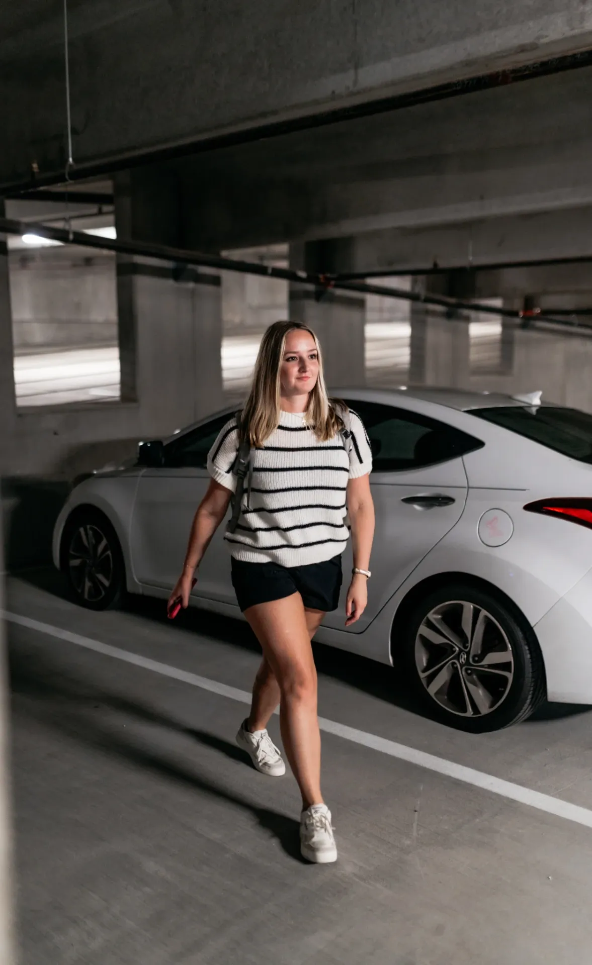 A woman with blonde hair, wearing a striped shirt, black shorts, and white sneakers, walks in a parking garage next to a white car. She holds a phone in her right hand and looks forward.