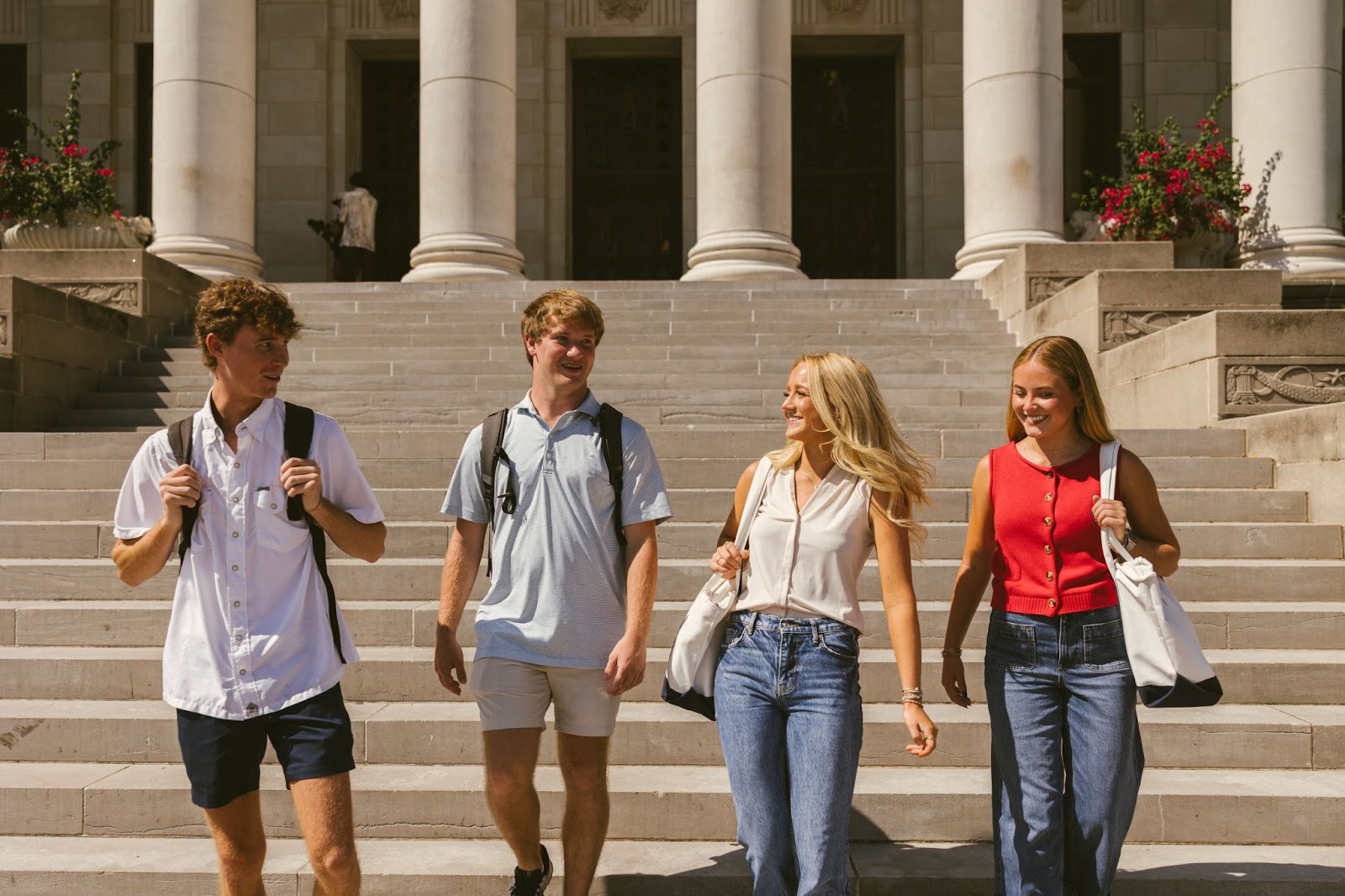 Four college students, two men and two women, walk down stone steps in front of a building with large columns—likely part of Texas A&M student housing. They are smiling, carrying bags, and enjoying a lively conversation together.