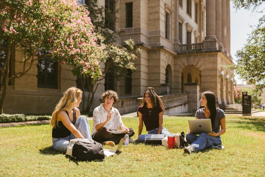 Four students sit on grass outside a large, historic building, chatting and smiling. Two have laptops; one holds a drink. With student apartments nearby and pink-flowered trees in view, it’s a relaxed campus study session.