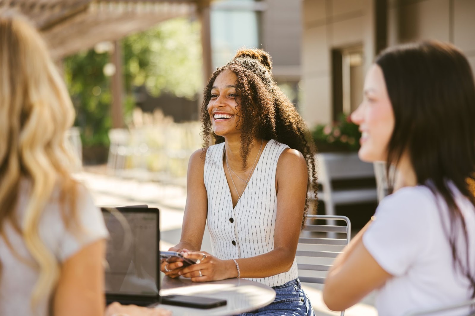 Three women sit together outside at a table, smiling and talking. One woman in the center, wearing a sleeveless shirt, holds a phone. There is a laptop on the table, and greenery is visible in the background.