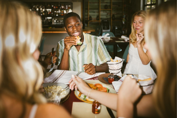 A group of friends sit around a restaurant table, eating and talking. One person in a green patterned shirt is smiling and taking a bite of food, while others are engaged and enjoying their meal.