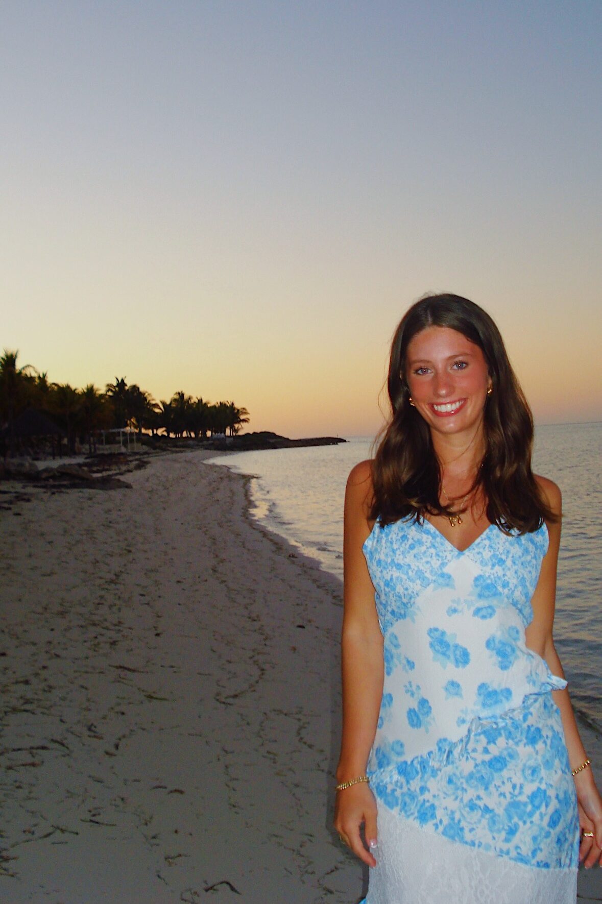 A woman in a blue and white floral dress smiles while standing on a sandy beach at sunset, dreaming of student apartment jobs in College Station, with calm ocean water and palm trees in the background.
