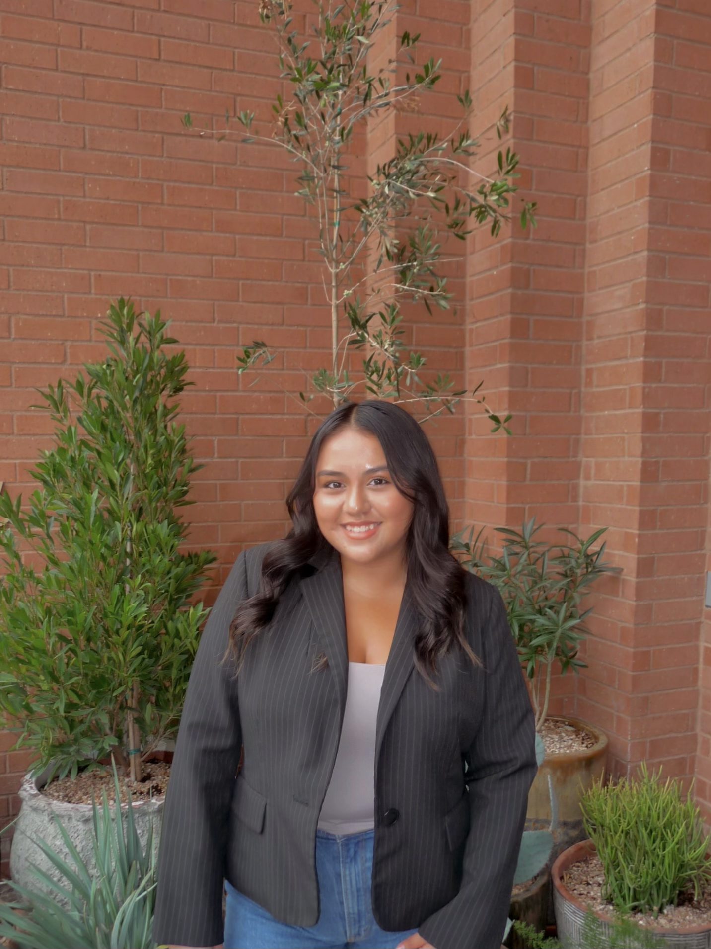 A woman with long dark hair, wearing a black pinstripe blazer and blue jeans, stands smiling in front of tall green potted plants and a brick wall—perfect for student apartment jobs in College Station.