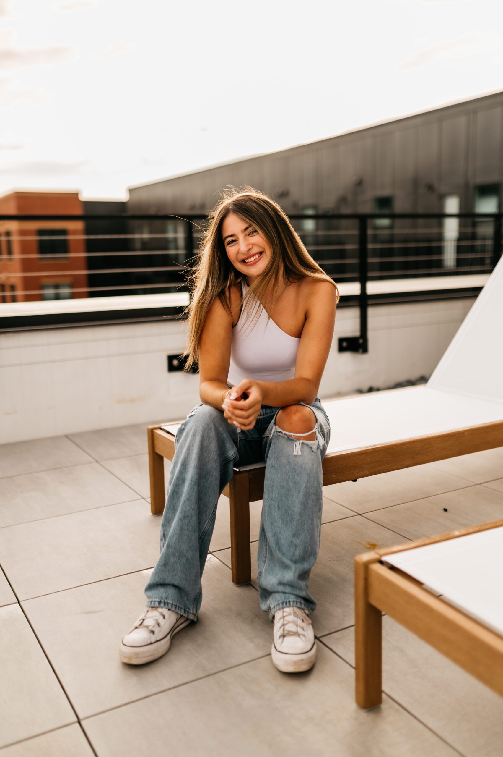 A young woman with long hair sits on a wooden lounge chair atop a student apartment in College Station, smiling. She wears a white one-shoulder top, ripped blue jeans, and white sneakers. The background shows buildings and a railing.
