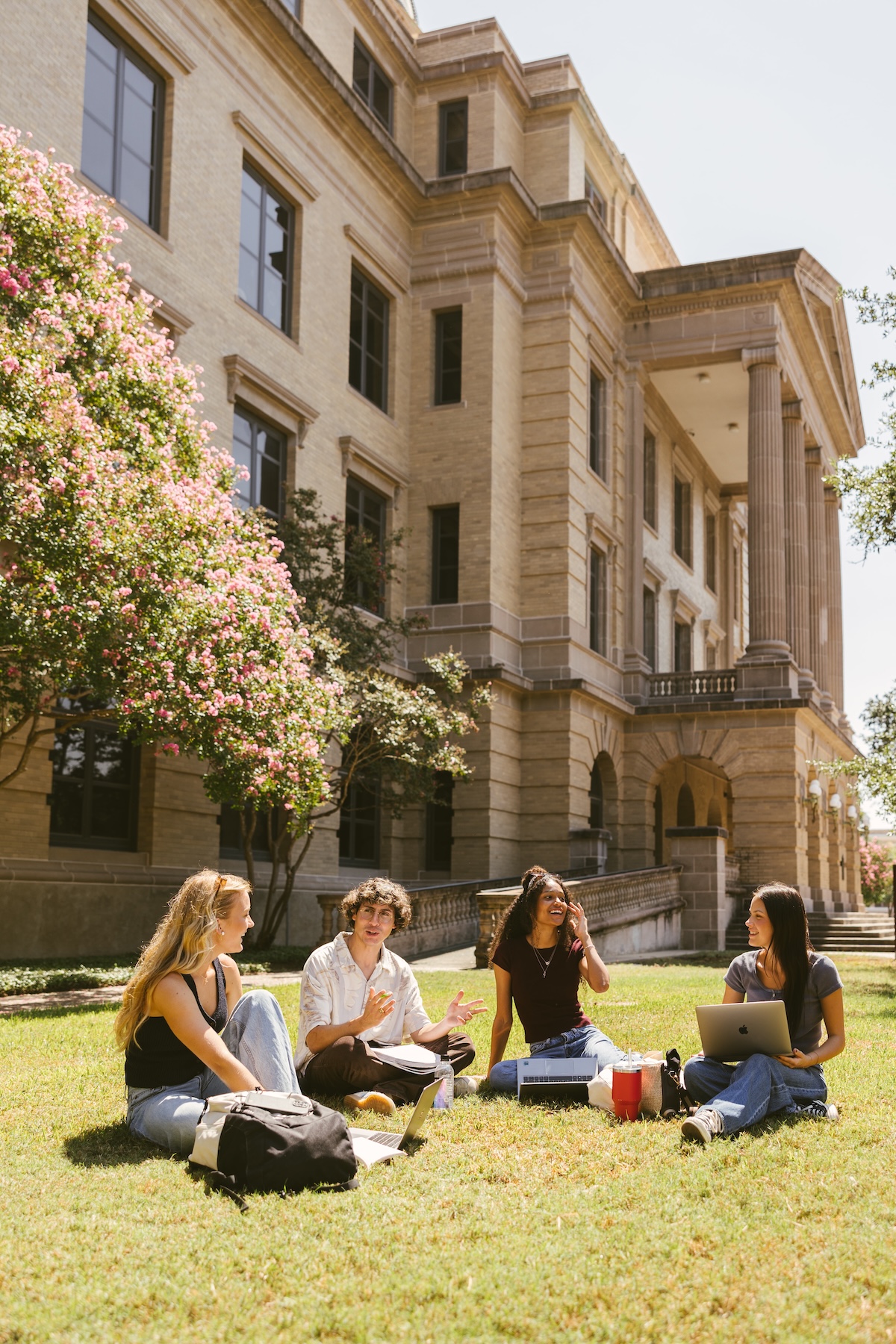 Four college students sit on grass outside a large academic building with columns in College Station, studying and chatting about student apartment jobs. Laptops, notebooks, and backpacks are spread around them. Trees with pink flowers frame the sunny scene.