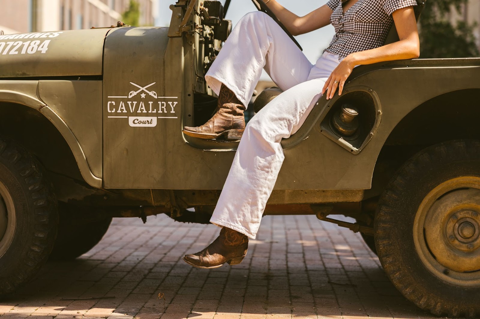 A person in white pants and cowboy boots sits on the open door of an olive-green Jeep labeled Cavalry Court, their lower body visible—capturing a classic scene outside Texas A&M parking.
