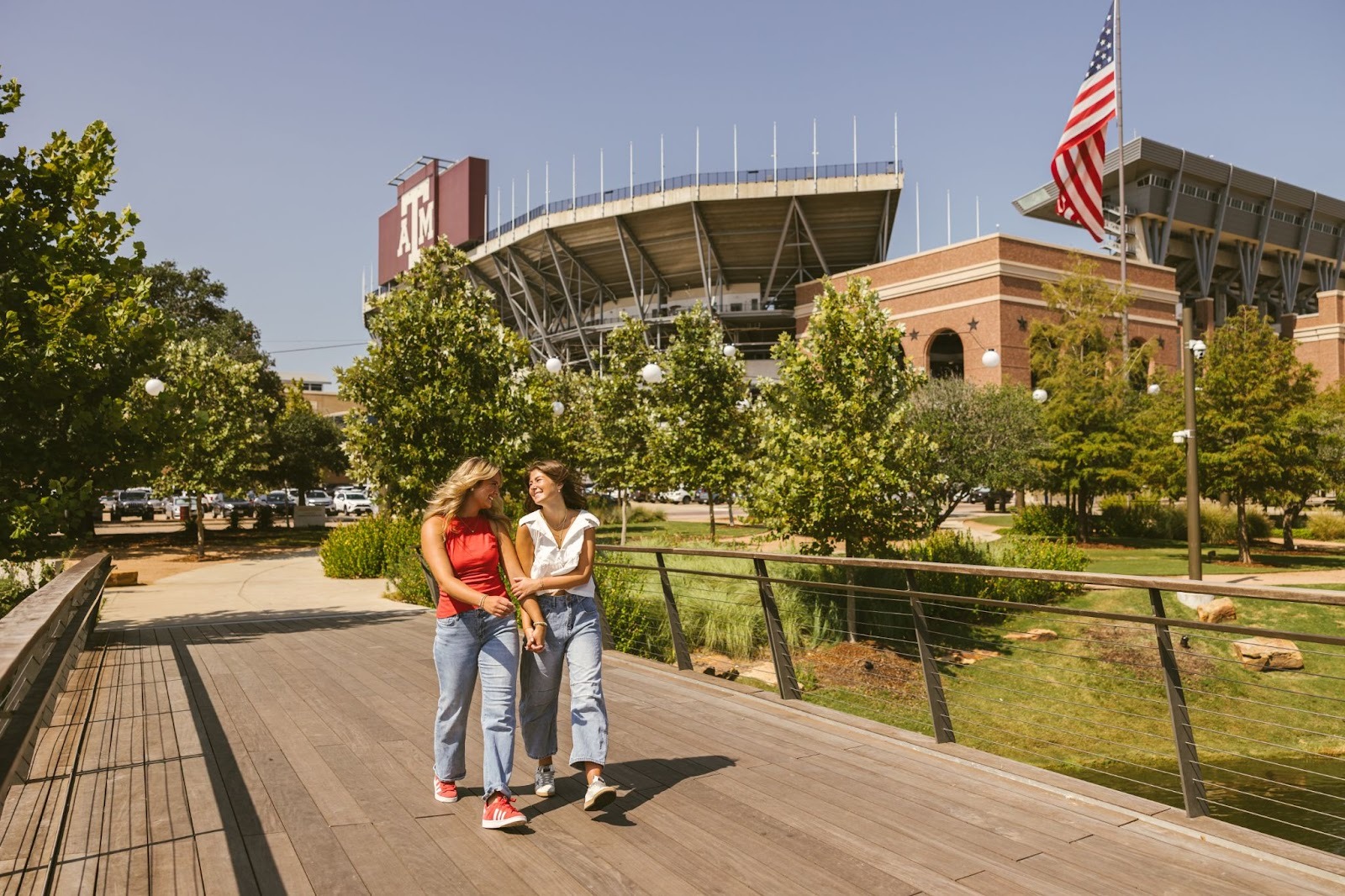 Two young women walk and smile on a wooden bridge surrounded by trees. Behind them is the Texas A&M University stadium, a large American flag, and nearby Texas A&M housing—all on a sunny day.