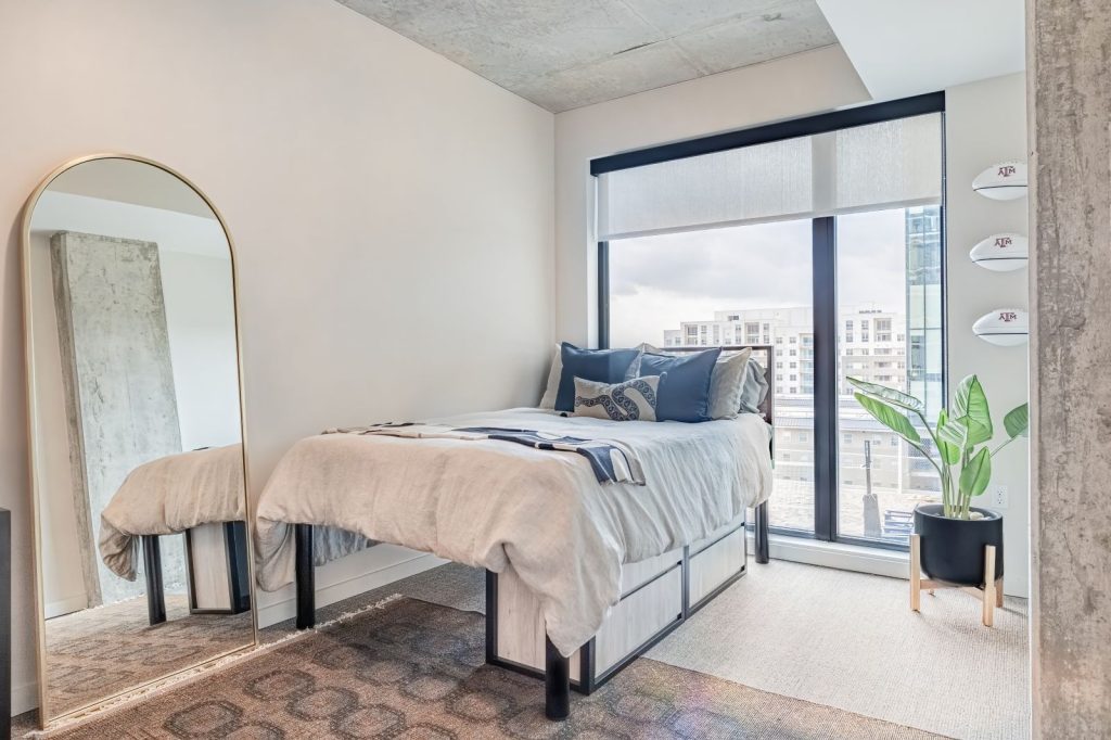A modern bedroom with a bed near large windows, a gold-framed floor mirror, a potted plant, and wall-mounted decor features neutral-toned furnishings and city views—perfect inspiration for stylish Texas A&M housing.