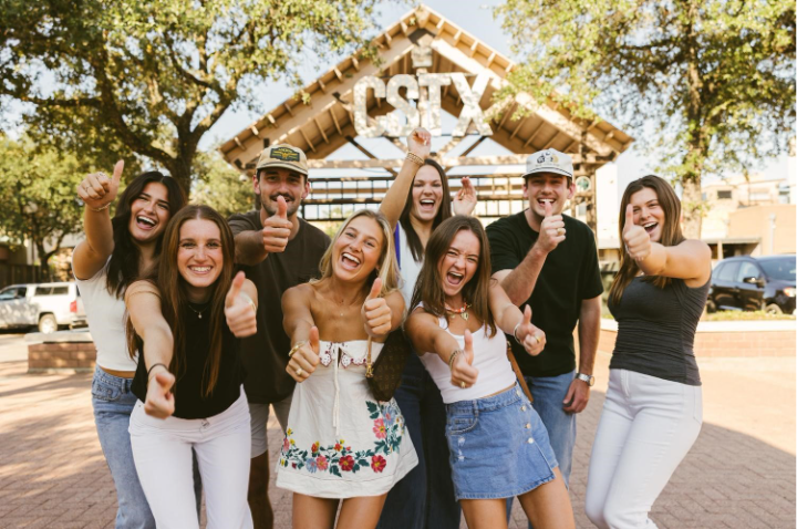 Texas A&M students huddled together, giving a thumbs up in front of theCollege Station Texas sign.