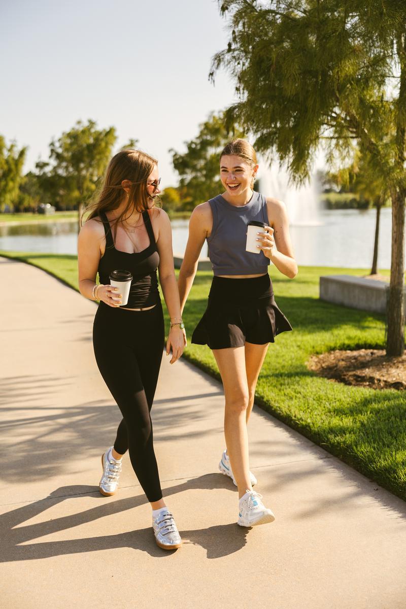 Two young women in athletic wear walk outdoors near a lake, holding coffee cups and smiling at each other. The scenic path by the fountain is just steps away from off campus apartments TAMU students love.