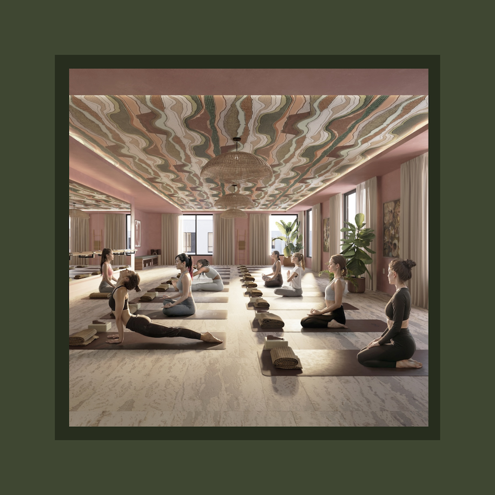 A group of people practice yoga in a spacious, sunlit studio with large windows, green plants, and a colorful wavy ceiling. They are seated on mats arranged in rows, following an instructor at the front.