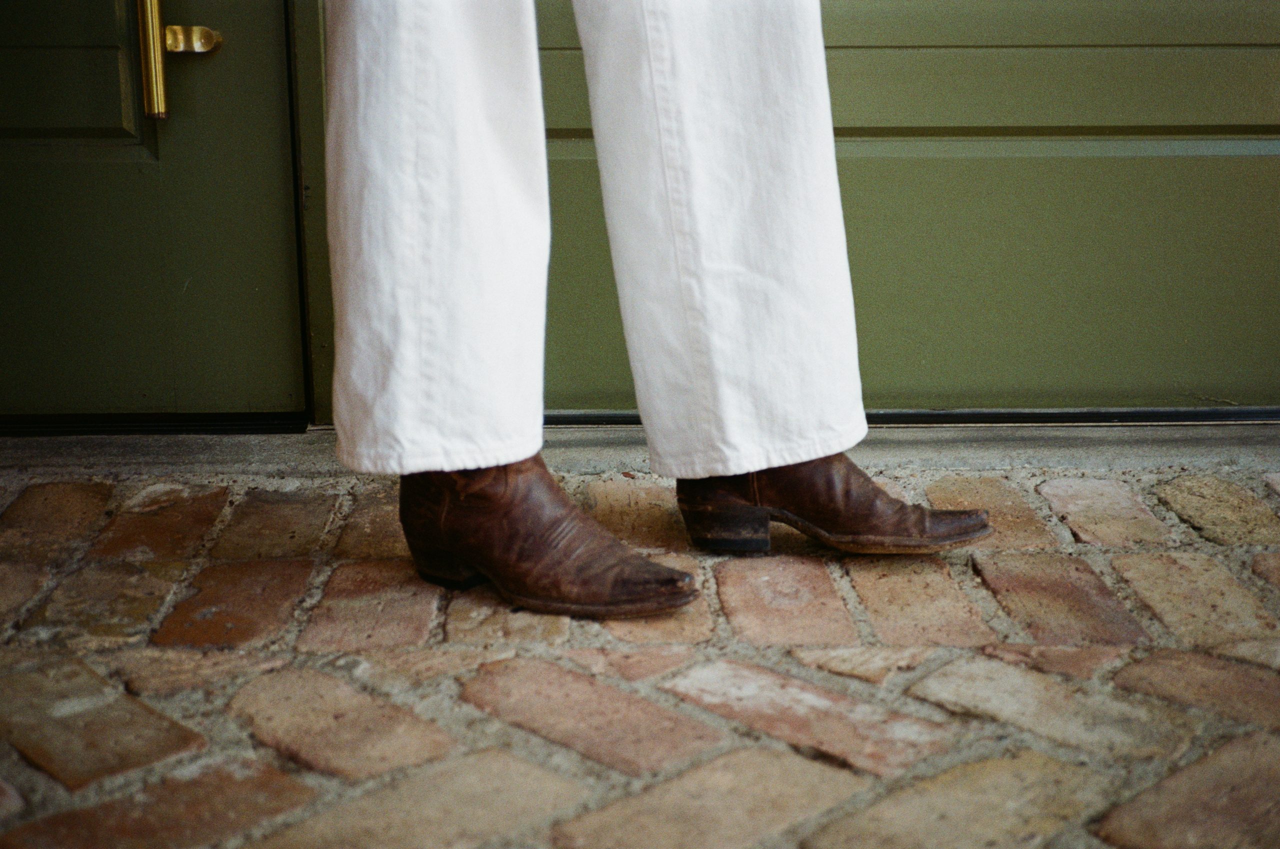 Person wearing white pants and brown cowboy boots stands on a brick floor in front of a green door.